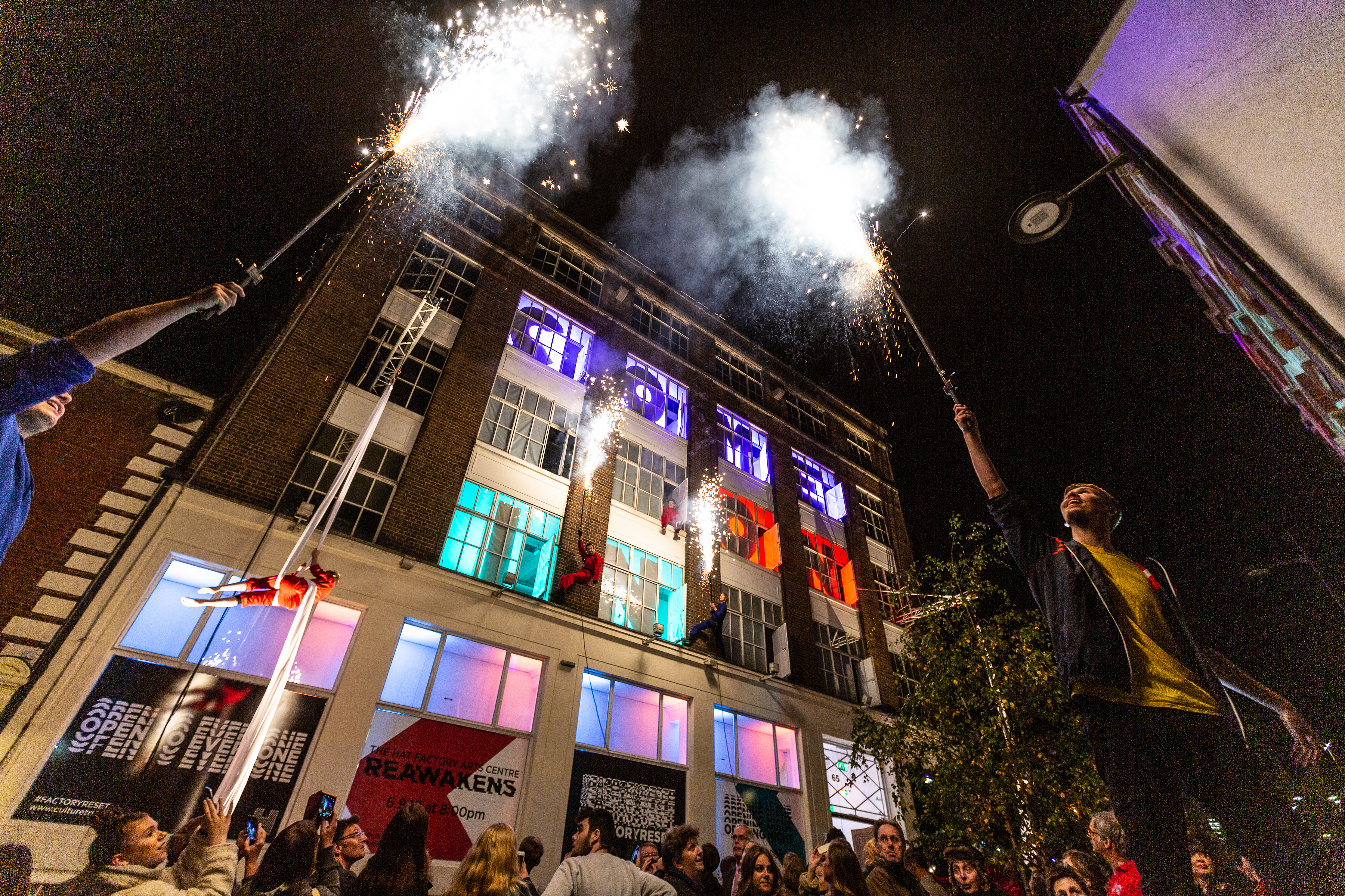 two performers hold giant sparklers in front of the hat factory, taken from factory reset