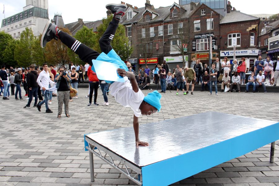 A young person wearing a blue hat and white t shirt, doing a one handed hand stand on a silver and blue plinth