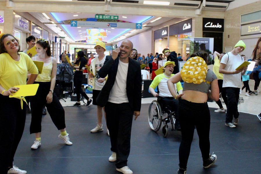 A man in a black suit and white t shirt on his phone surrounded by young people moving in a flurry around him (wearing yellow themed costume) in a shopping centre