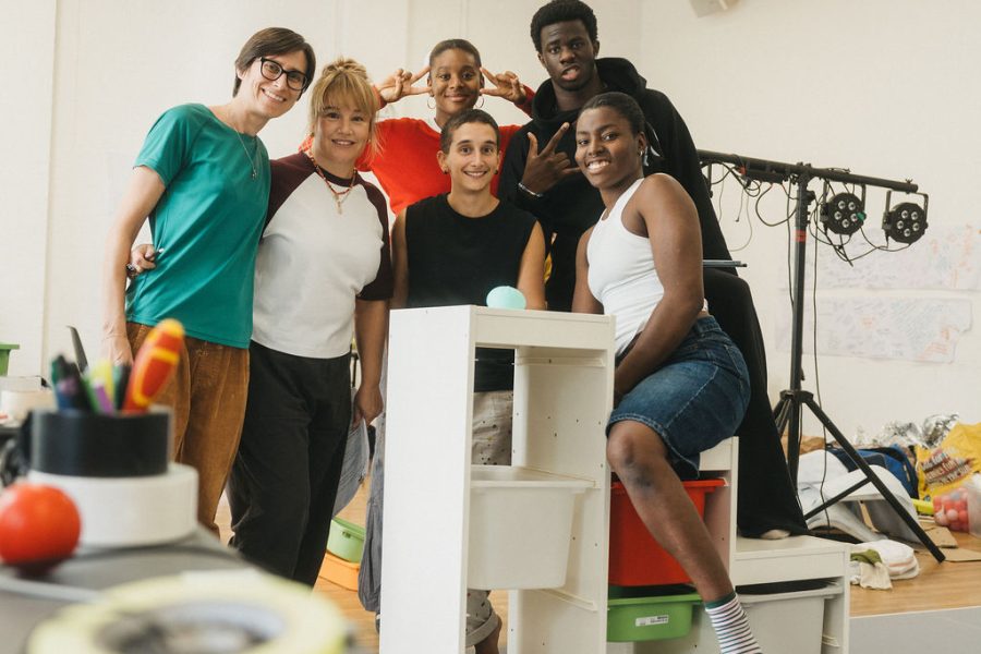 A rehearsal shot of the cast of A Class of Your Own looking directly at the camera on the set, with rehearsal props in the foreground out of focus.