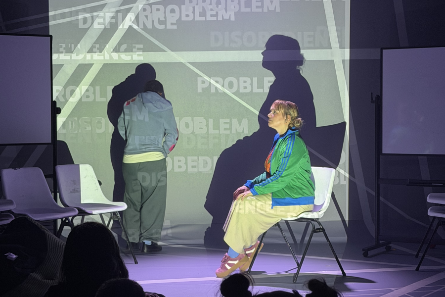 Emily (Deb) sits on a chair in profile onstage, their shadow behind them. Another actor stands with back to audience. There are back of heads of a schools audience in forefornt.