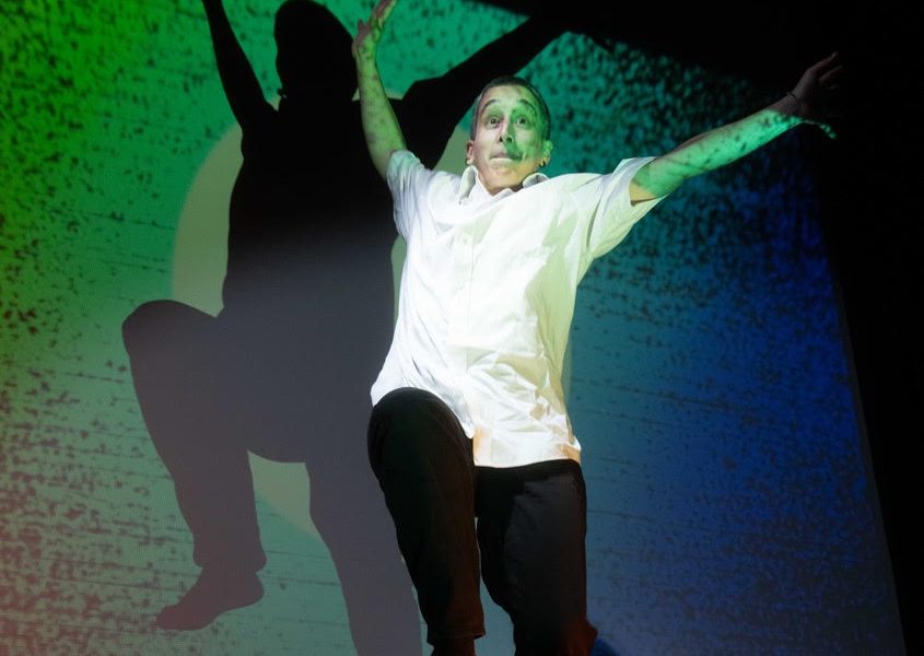 A performer balancing on one leg on top of a chair with arms outstretched with a green lit background and shadow of the performer on the background