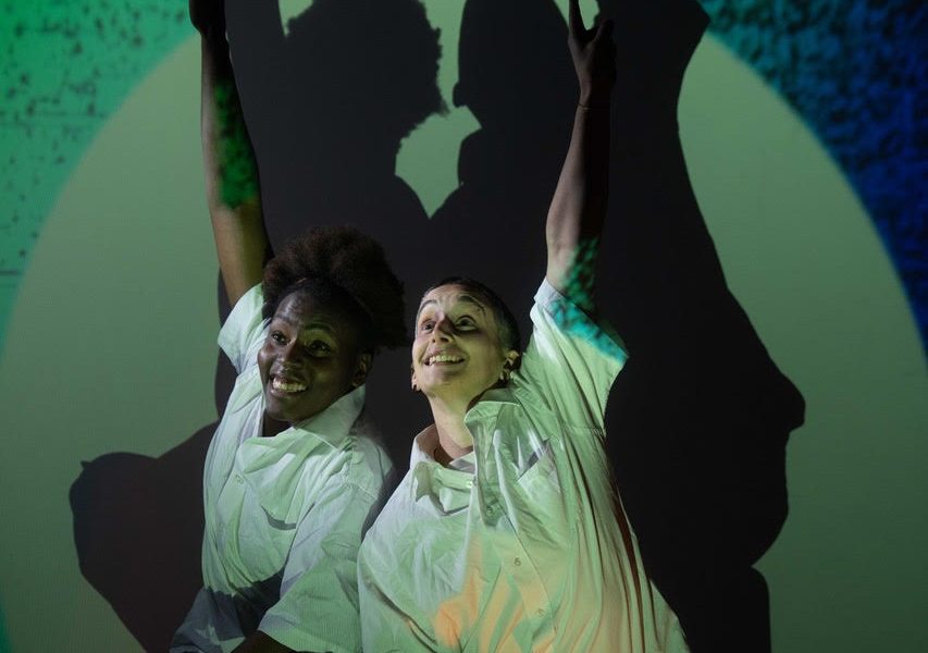 Two actors reaching their hands up as of to answer a question with smiling faces, against a Green background and spotlight with shadows of the performers behind.
